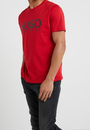 Red cotton t-shirt with short sleeves featuring a black "HUGO" logo across the front. Worn with dark gray jeans.