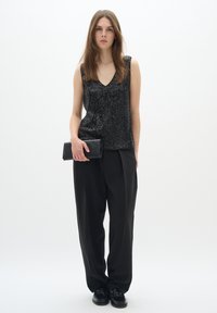 Top senza maniche in paillettes nere con scollo a V, abbinato a pantaloni neri larghi e una pochette nera. Texture liscia, design minimalista.
