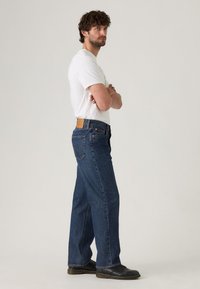 Jeans de mezclilla azul con un corte recto, que presentan una parche de cuero en la parte trasera y bolsillos traseros dobles. Combinados con una camiseta blanca básica y zapatos negros.