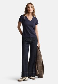 Tricou bleumarin din bumbac cu decolteu în V, croială lejeră, asortat cu pantaloni din denim închiși, evazați, și adidași bej. Un sacou în carouri este așezat pe un braț.