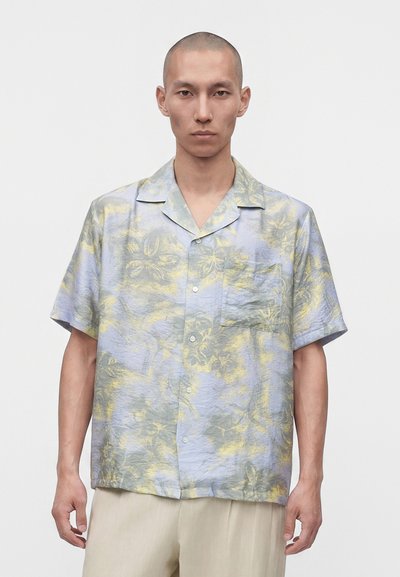 Hombre con cabeza rapada que lleva una camisa de botón con mangas cortas, de color azul pastel y amarillo con flores, y pantalones beige claro sobre fondo blanco.