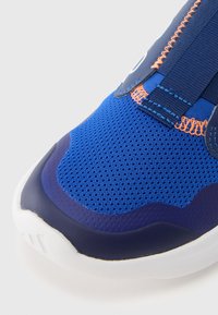 Zapatilla deportiva azul con material de malla transpirable, que presenta un patrón texturizado, correas elásticas y suela de goma duradera en blanco contrastante.