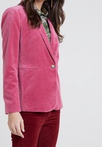 Růžový sametový blazer s jedním knoflíkem, špičatými klopami a bočními kapsami, ve spojení s červenými sametovými kalhotami, které ukazují hladkou texturu.