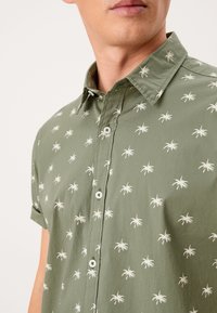 Chemise à manches courtes vert olive avec motif de palmiers blancs, dotée d'un col boutonné et de boutons sur le devant, fabriquée en tissu léger.