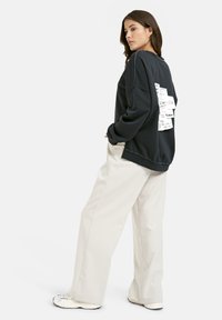 Frau trägt einen schwarzen Hoodie mit weißen Textpatches auf dem Rücken, weite beige Hose und weiße Sneaker, steht seitlich mit den Händen in den Taschen.