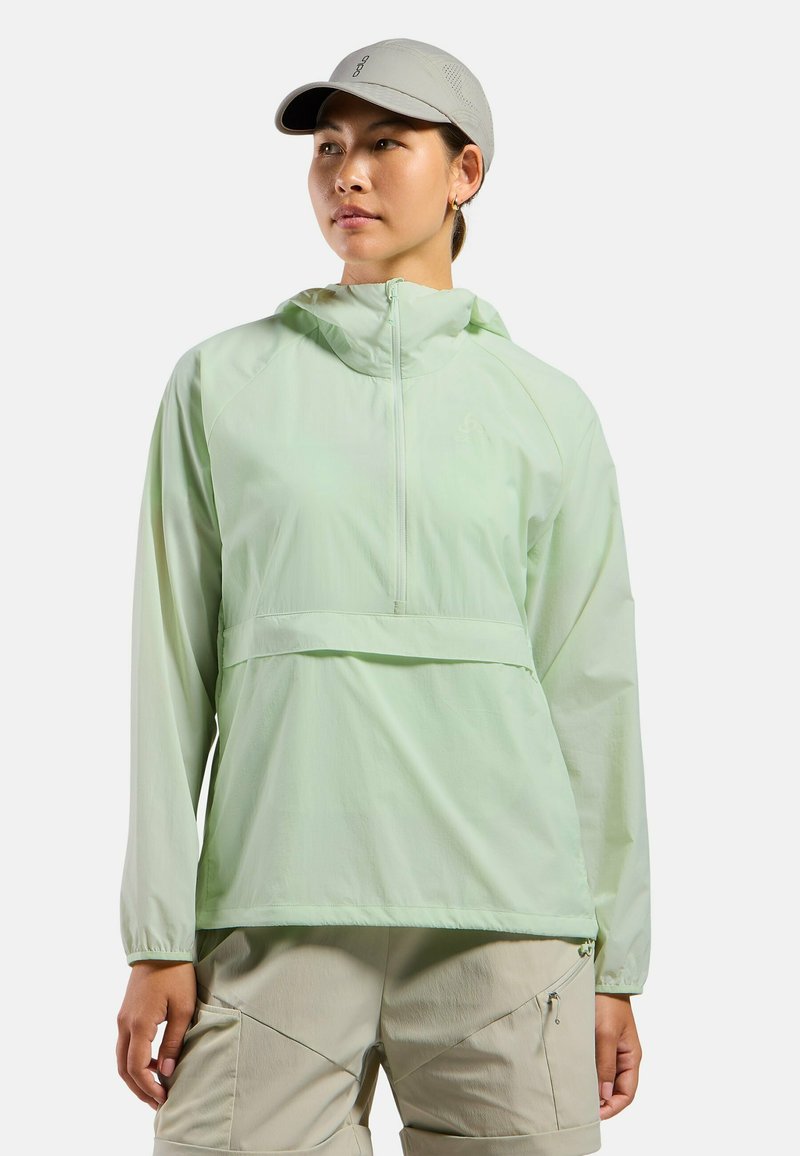 Leichtgrüner, leichter Pulli-Jacke mit Kapuze, halbem Reißverschluss vorne und Kängurutasche; kombiniert mit hellbeigen Cargo-Hosen.