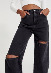 Personne portant un jean noir déchiré taille haute avec de grandes trous aux genoux et un top blanc court à manches longues, les mains dans les poches.