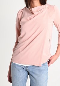 Woman wearing a light pink long-sleeve top with an asymmetrical button detail and zip pockets, paired with light blue jeans.