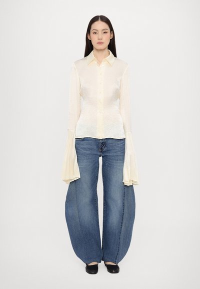 Camicia button-up di un giallo chiaro, con una texture, maniche lunghe e svasate abbinata a jeans in denim blu a gamba larga. Scarpe nere completano l'outfit.