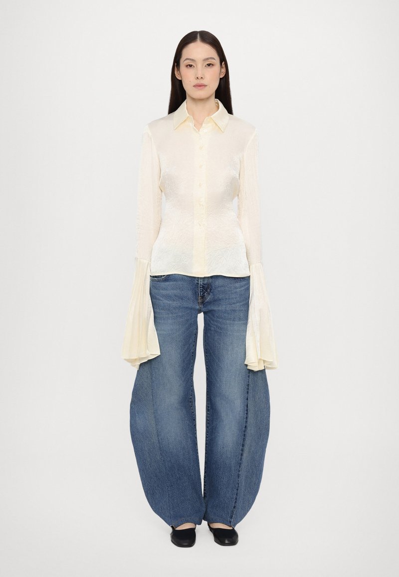 Cămașă călcată, de un galben deschis, cu textura, cu mâneci lungi evazate, purtată împreună cu blugi albaștri cu picioare largi. Pantofi negri completează ținuta.