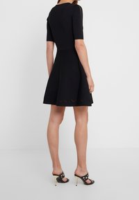 Abito nero a maniche corte con corpino aderente e gonna svasata, caratterizzato da un dettaglio a zigzag ritagliato sull'orlo. La modella indossa sandali neri con tacco.