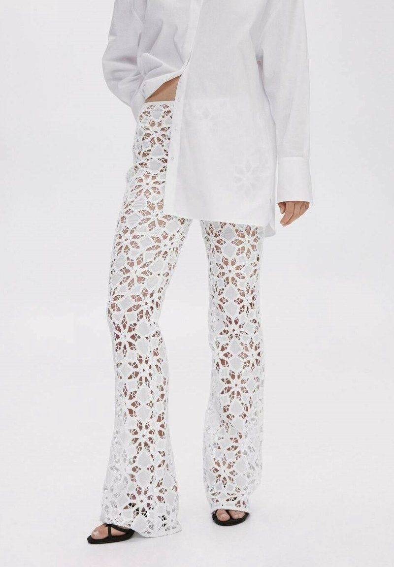 Femme portant un pantalon en dentelle florale blanche et une longue chemise blanche boutonnée avec des sandales noires à bouts ouverts sur un fond uni.
