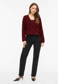 Bordo spalvos megztinis su užmovimu, minkšta tekstūra ir ilgomis rankovėmis, derinamas su juodais aukštakulniais džinsais ir juodais bateliais su smailia nosimi.