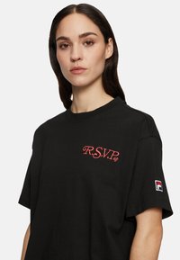 Camiseta de algodón negra con logotipo en cursiva rojo "R.S.V.P" en el pecho y etiqueta de la marca FILA en la manga. Mangas cortas y ajuste relajado.