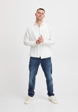 Biała lniana koszula na guziki z kieszenią na piersi, zestawiona z niebieskimi dżinsami i białymi trampkami, prezentowana na stojącym męskim modelu.