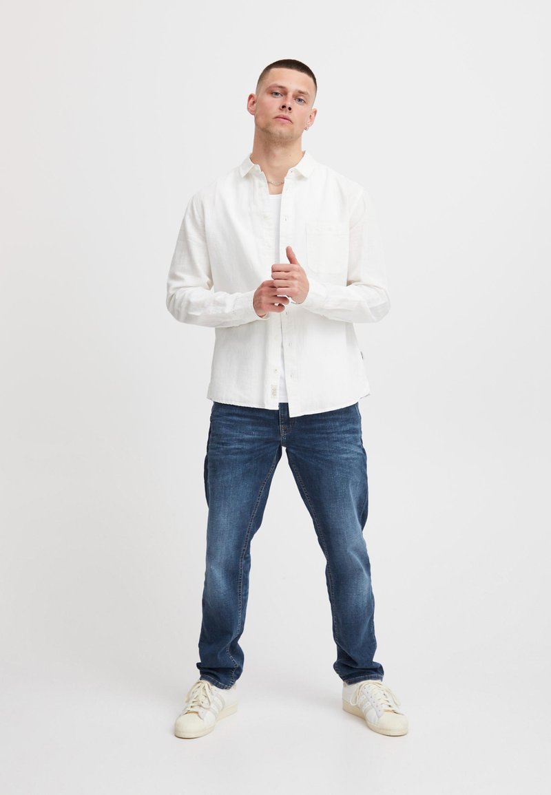 Hvit skjorte i lin med knapper og brystlomme, kombinert med blå denimjeans og hvite sneakers, vist på en stående mannlig modell.