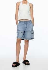 Haut dos nu gris clair, short cargo en jean bleu taille haute et ajusté, avec deux poches latérales, porté avec des claquettes noires nouées.