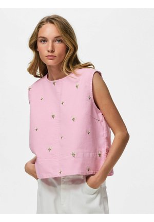 Femme aux cheveux blonds porte un haut rose sans manches avec de petites fleurs blanches imprimées et un pantalon blanc, debout les mains dans les poches.