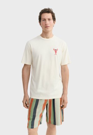 Mann trägt ein cremefarbenes T-Shirt mit einem kleinen roten Hummer-Design auf der Brust und gestreifte Shorts in Grün, Orange und Marineblau.