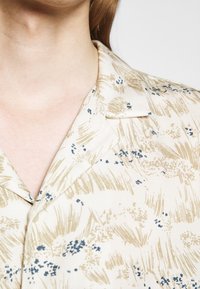 Camicetta a maniche corte in tessuto crema con motivi floreali astratti in beige e blu, dotata di colletto classico e texture liscia.