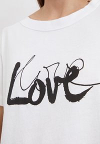 Weißes Baumwoll-T-Shirt mit fett gedrucktem, handgeschriebenem Text, der "Liebe" buchstabiert. Der Stoff hat eine weiche Textur und einen klassischen Rundhalsausschnitt.