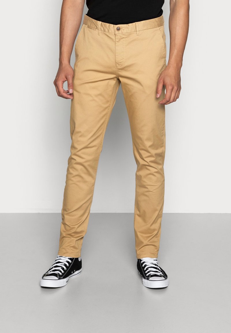 Beige chinos med slim fit, lavet af bomuldsblandingsstof. Har frontlommer, knaplukning og bæres med sort-hvide sneakers.