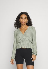 Top verde com mangas longas, padrão texturizado e bainha peplum, combinado com calções de bicicleta pretos. Tecido escovado, acabamento suave.