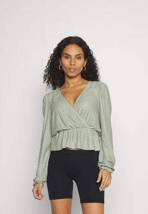 Top verde com mangas longas, padrão texturizado e bainha peplum, combinado com calções de bicicleta pretos. Tecido escovado, acabamento suave.