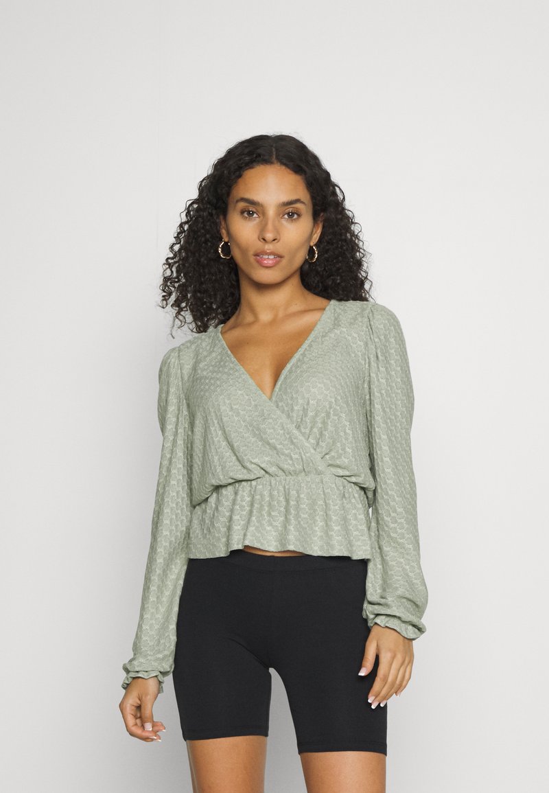 Top verde com mangas longas, padrão texturizado e bainha peplum, combinado com calções de bicicleta pretos. Tecido escovado, acabamento suave.