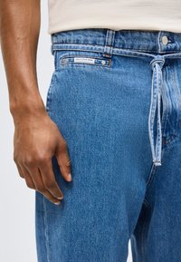 Uomo che indossa jeans in denim azzurro chiaro con una cintura in tessuto annodata, e un'etichetta bianca sulla tasca in vita con la scritta "Marc O'Polo Denim".