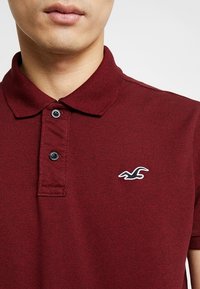 Mörkröd pikétröja i texturerat tyg med krage, två svarta knappar och en vit fågel-logotyp på vänster bröst.