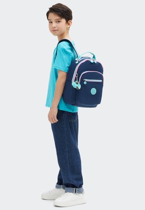 Ragazzo giovane con una maglietta turchese, jeans scuri, sneakers bianche, che porta uno zaino blu navy con dettagli azzurri e rosa, rivolto di lato.