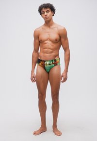 Νεαρός άνδρας με σγουρά μαλλιά, όρθιος, ξυπόλητος, φορώντας πολύχρωμο μαγιό με σχέδια, μπροστά σε απλό ανοιχτό γκρι φόντο.