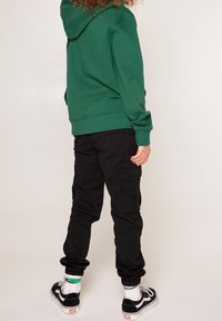Zelená kapucňová mikina, černé cargo kalhoty a černé tenisky s bílými akcenty. Viditelná textura na látce mikiny a kapsách kalhot.