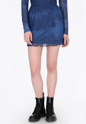 Vrouw draagt een doorschijnende marineblauwe mini-rok en een top met lange mouwen, gecombineerd met zwarte leren veterlaarzen, staand tegen een witte achtergrond.