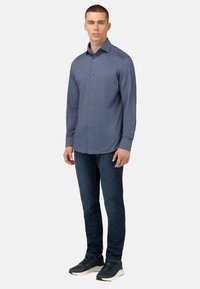 Šviesiai mėlynas ilgas rankovėmis marškinis su mygtukų užsegimu, derinamas su tamsiai mėlynomis džinsais ir pilkais batais, stovintis ant vienodo fono.