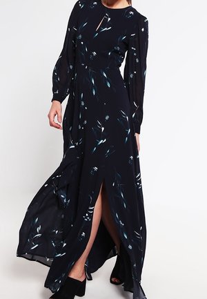 Kobieta ubrana w długą czarną sukienkę w kwiaty z długimi rękawami i rozcięciem z przodu, zestawioną z czarnymi sandałami na blokowym obcasie z odkrytymi palcami.