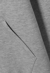 Pilka medžiaga, turinti lygią tekstūrą ir subtilų margumą. Komplekte yra pasviręs priekinis kišenėlis su švariais siūlių detalėmis.