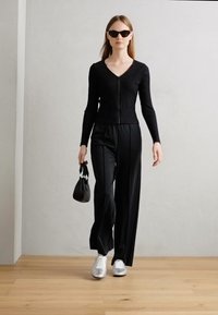 Haut noir à manches longues côtelé avec une fermeture éclair à l'avant, associé à un pantalon noir à larges jambes. Porté avec des baskets argentées et un petit sac à main noir.