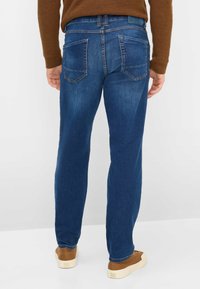 Jeans in denim blu scuro con vestibilità dritta, caratterizzati da due tasche posteriori e cuciture a contrasto, indossati con scarpe marroni e un maglione marrone.