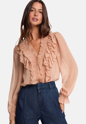 Mujer con una blusa transparente de color rosa pálido con volantes, de manga larga y vaqueros azul oscuro de talle alto, de pie frente a un fondo blanco.