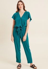 Tutina teal con scollatura a V, maniche a farfalla, vita con cintura e gambe affusolate. Materiale liscio con una sottile texture. Sandali neri.