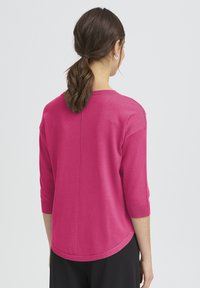 Pull rose vif à manches trois-quarts, col arrondi et ourlet légèrement incurvé. Texture en maille avec une finition lisse.