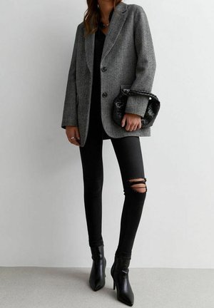 Person iført en grå blazer, sorte revnede skinny jeans, sorte ankelstøvler med hæl, der holder en lille sort flettet håndtaske.