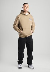 Beige hoodie with a front pocket and "USA" logo, paired with black pants. Model wears white sneakers. Simple, casual design.