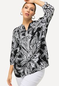 Sort-hvid blomstret blouse med V-udskæring, trekvartærmer og asymmetrisk kant. Stoffet har en glat, let tekstur.