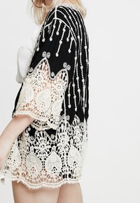 Kimono in pizzo nero con dettagli in crochet bianco, motivi a cuori e perline decorative lungo le maniche e l'orlo. Design con apertura frontale.