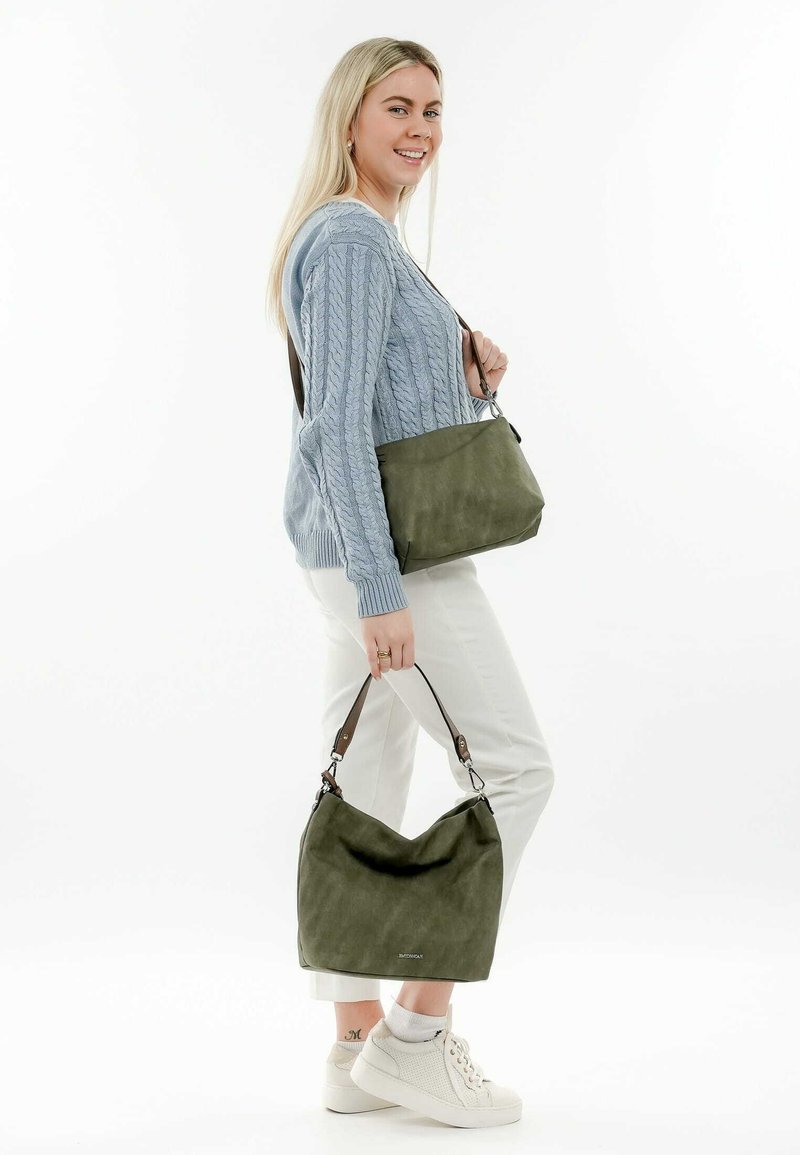 Olivgrüne Tasche mit glatter Textur, ausgestattet mit kontrastierenden braunen Ledergriffen. Robuste Hobo-Form, getragen von einem Model in lässiger Kleidung.
