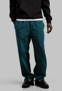 Turkusowe spodnie typu wide-leg uszyte z miękkiego materiału, z plisowanym przodem, w połączeniu z czarną bluzą z kapturem i czarnymi mokasynami.