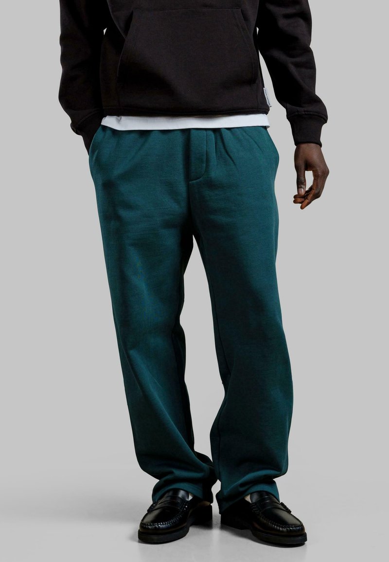 Turkusowe spodnie typu wide-leg uszyte z miękkiego materiału, z plisowanym przodem, w połączeniu z czarną bluzą z kapturem i czarnymi mokasynami.